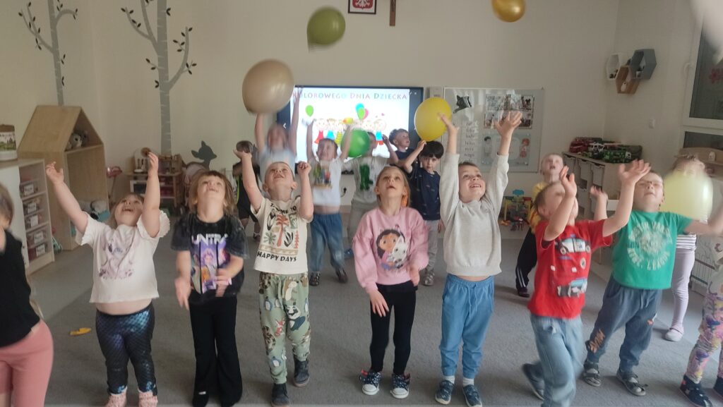 Grupa dzieci stoi na szarym dywanie i podrzuca do góry kolorowe balony.