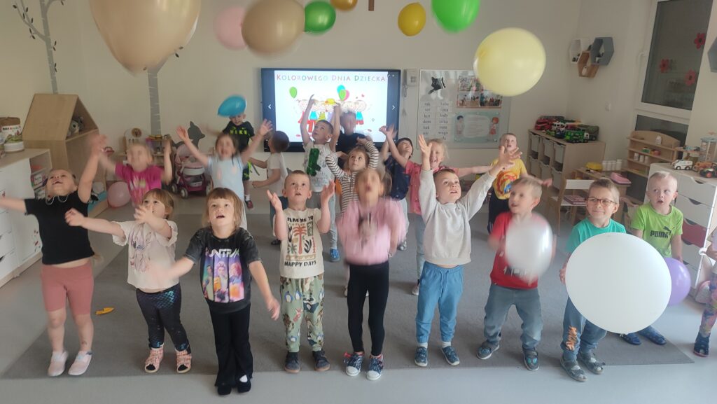Grupa dzieci stoi na szarym dywanie i podrzuca do góry kolorowe balony.