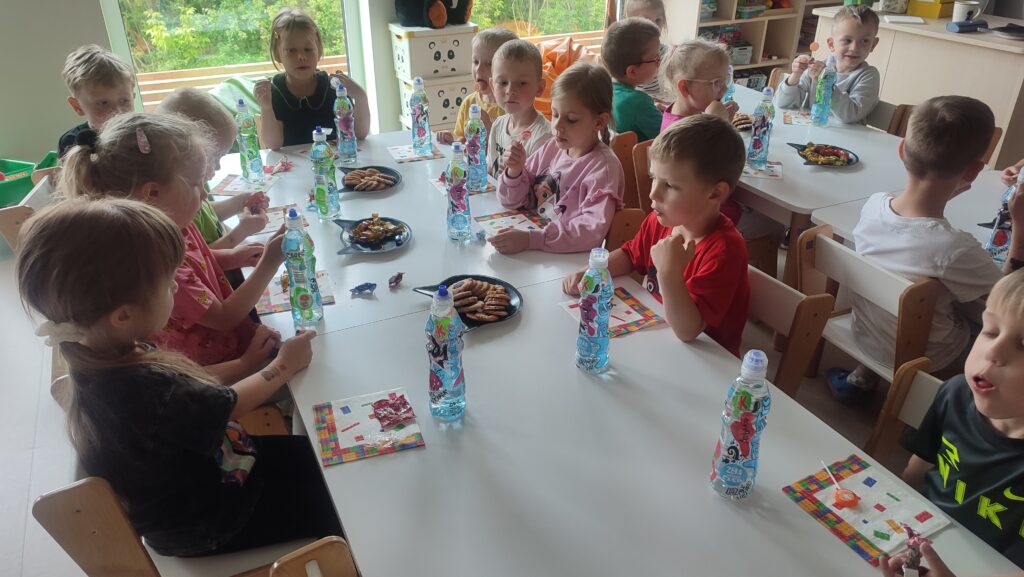 Dzieci siedzą przy białych stolikach, liżą lizaki i piją soki. Na stole kolorowe serwetki.