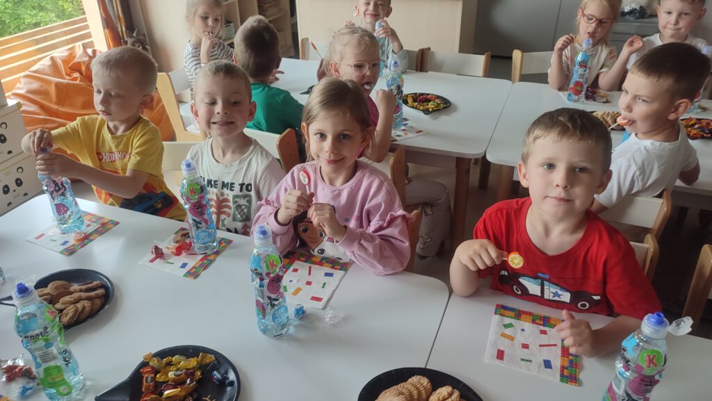 Dzieci siedzą przy białych stolikach, liżą lizaki i piją soki. Na stole kolorowe serwetki.