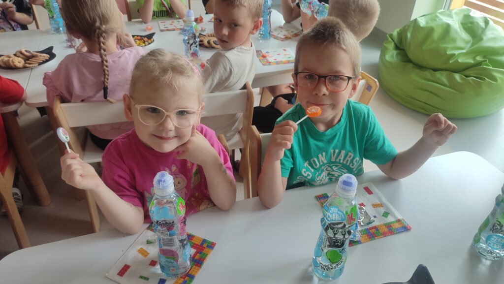 Dzieci siedzą przy białych stolikach, liżą lizaki i piją soki. Na stole kolorowe serwetki.