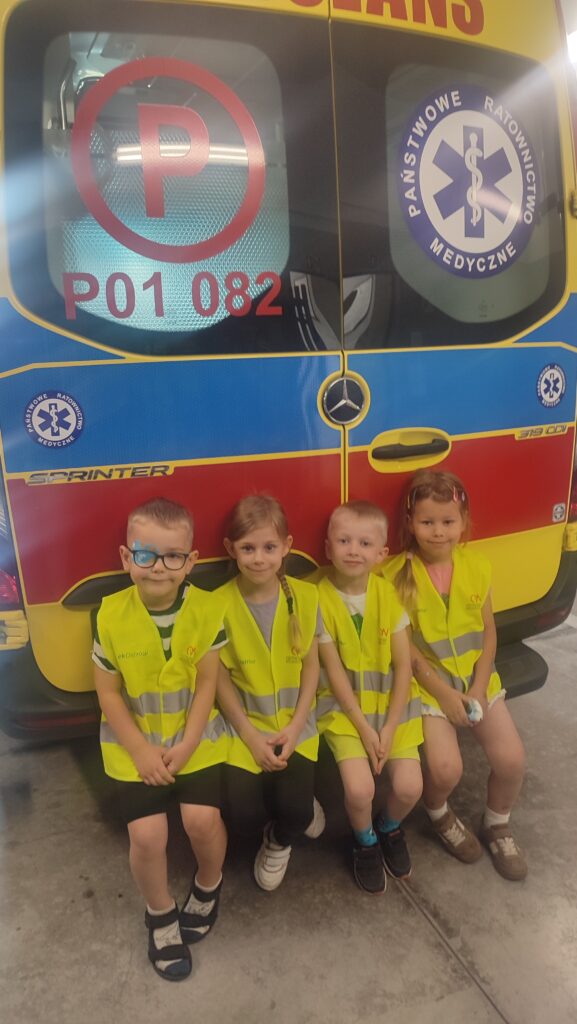 Czworo dzieci w żółtych kamizelkach siedzi na stopniach karetki pogotowia.