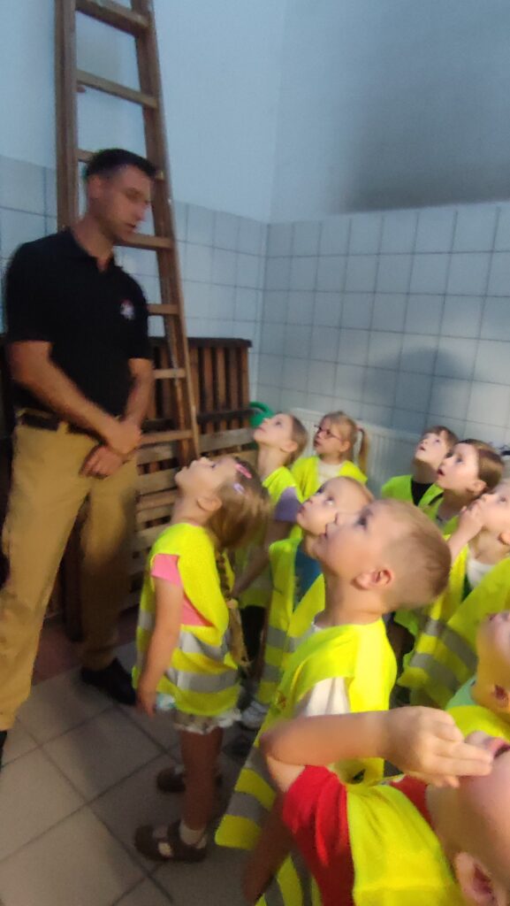 Dzieci w żółtych kamizelkach stoi razem ze strażakiem i spogląda do góry.