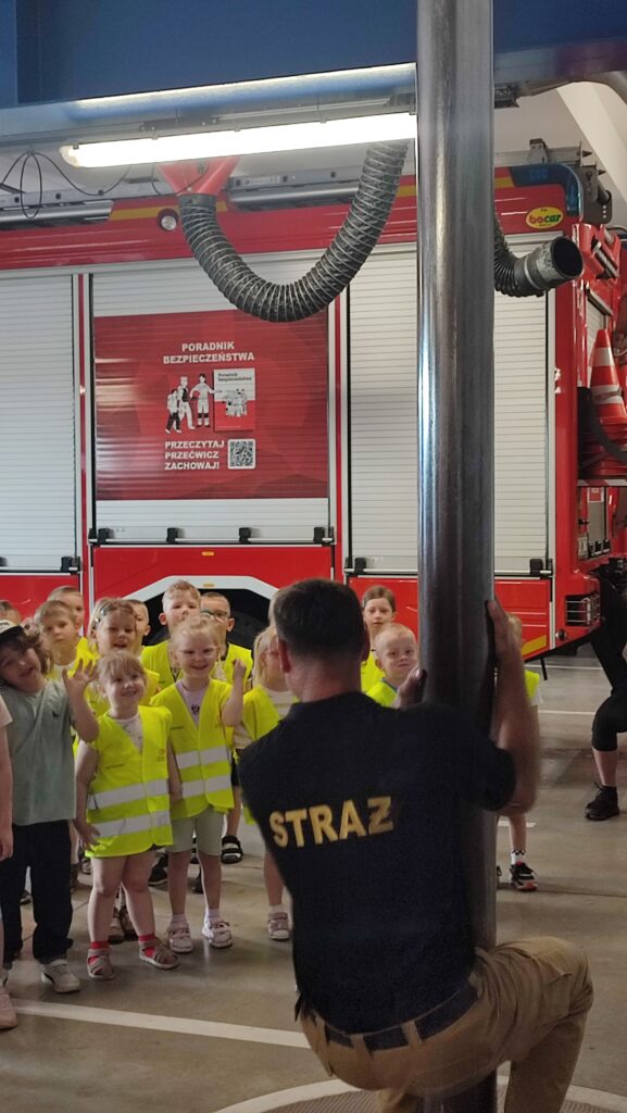 Grupa dzieci obserwuje zjeżdżającego strażaka z rury.