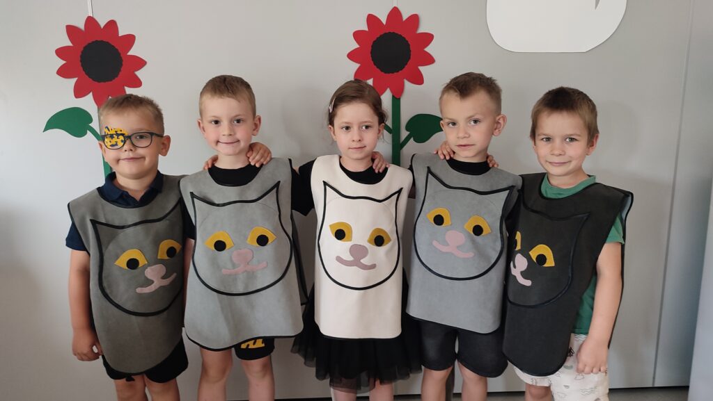Pięcioro dzieci ubranych na czarno w kocich kamizelkach pozuje do wspólnego zdjęcia na tle szarej ściany z czerwonymi kwiatkami.