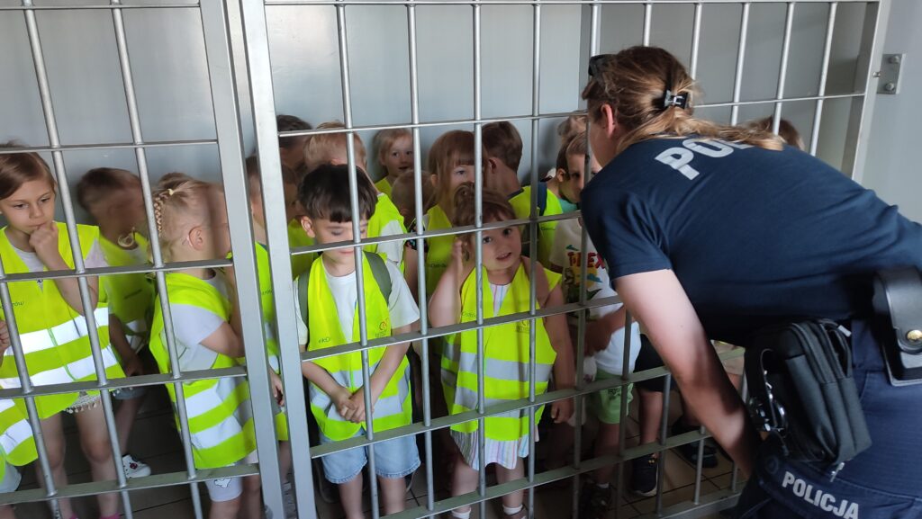 Duża grupa dzieci jest zamknięta za kratami. Przed nimi stoi policjantka.