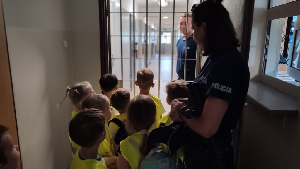 Dzieci w żółtych kamizelkach stoją przed kratami, za którym jest długi korytarz. Przy kracie stoją policjanci.