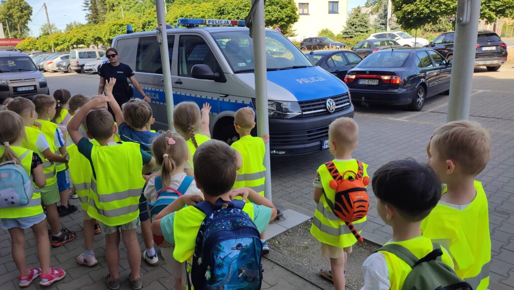 Duża grupa dzieci stoi na chodniku i patrzy na policjantkę i samochód policyjny.