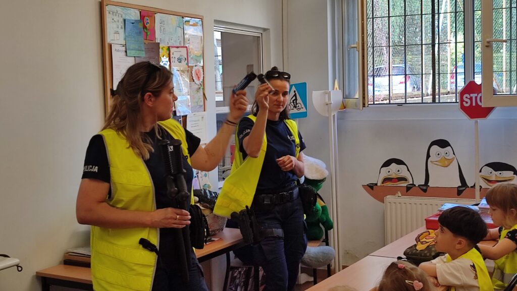 Dwie policjantki w granatowych mundurach pokazują dzieciom sprzęt używany przez policję.