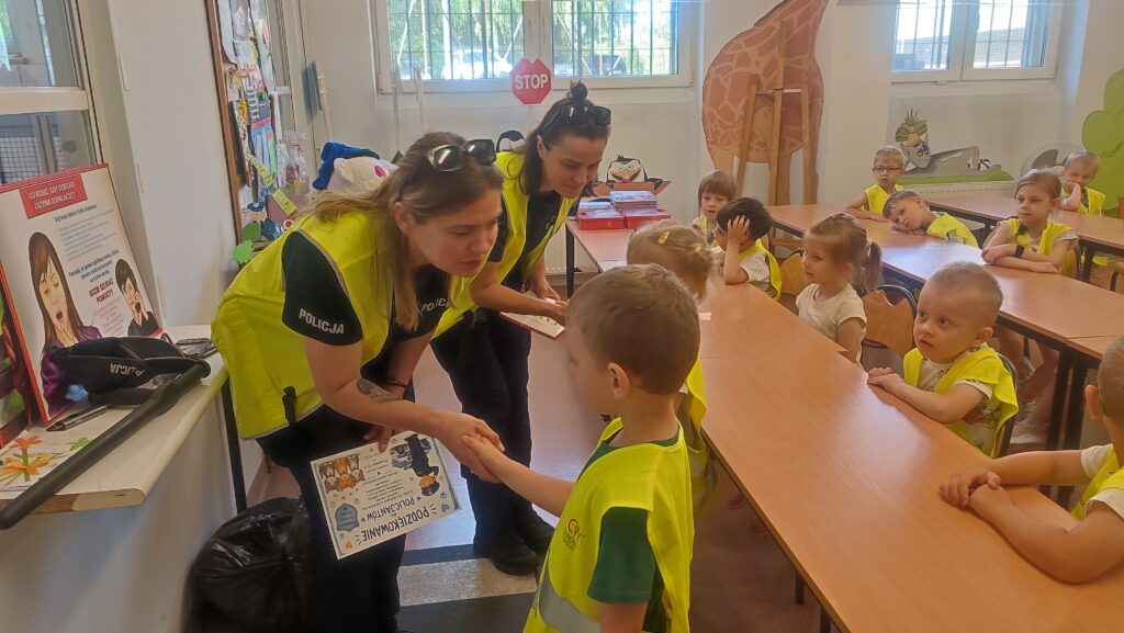 Chłopiec i dziewczynka w żółtych kamizelkach wręczają prezent policjantkom.