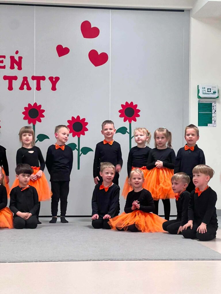 Duża grupa dzieci ubrana na czarno z dodatkami pomarańczowymi stoi na szarym dywanie na tle ściany udekorowanej w czerwone kwiaty.