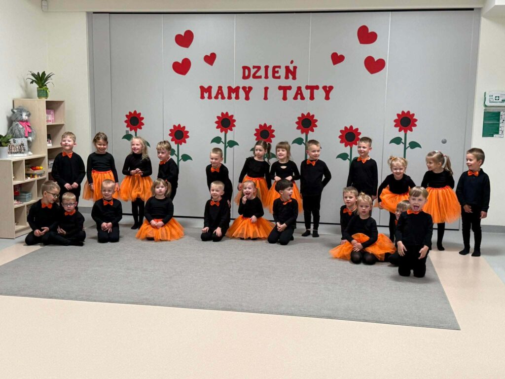 Duża grupa dzieci ubrana na czarno z dodatkami pomarańczowymi stoi na szarym dywanie na tle ściany udekorowanej w czerwone kwiaty.