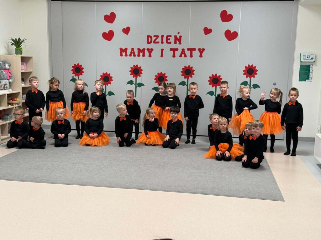 Duża grupa dzieci ubrana na czarno z dodatkami pomarańczowymi stoi na szarym dywanie na tle ściany udekorowanej w czerwone kwiaty.