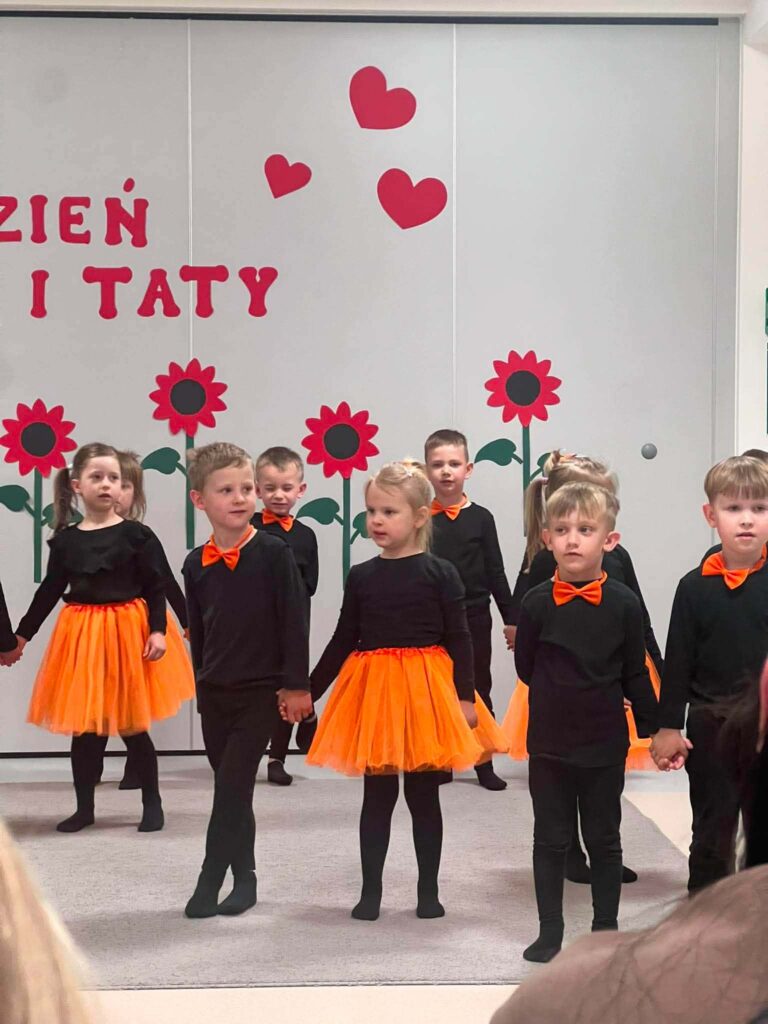 Duża grupa dzieci ubrana na czarno z dodatkami pomarańczowymi stoi na szarym dywanie na tle ściany udekorowanej w czerwone kwiaty.