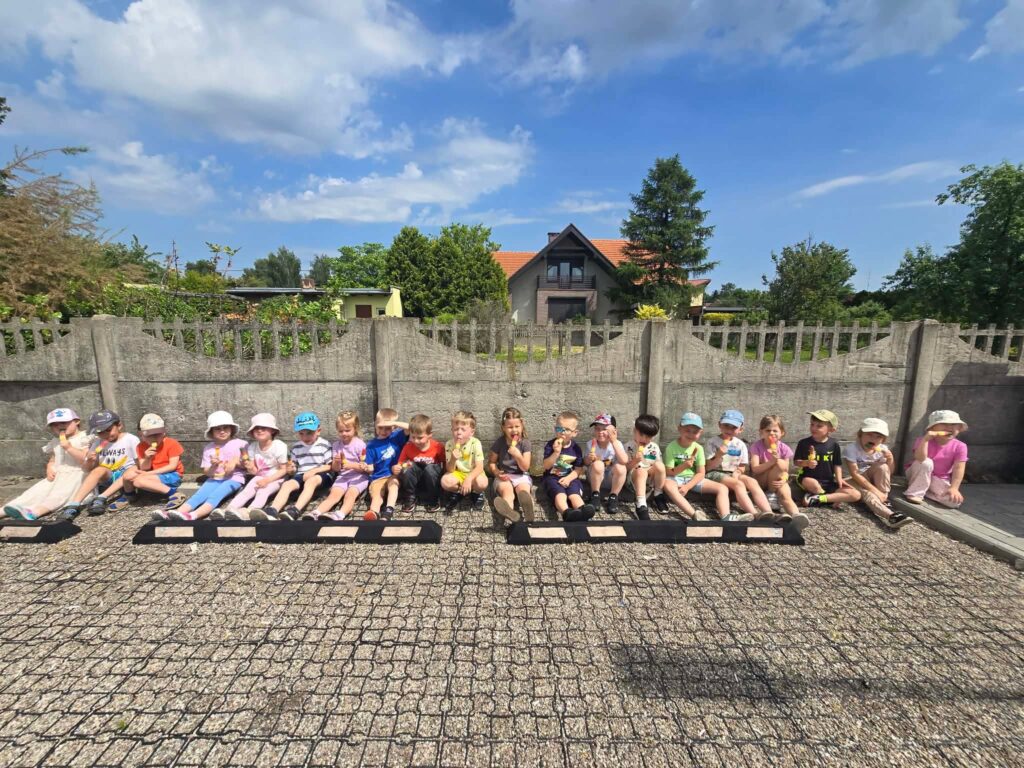 Duża grupa dzieci ubranych w krótkie spodenki siedzi pod betonowym płotem i liże lody.