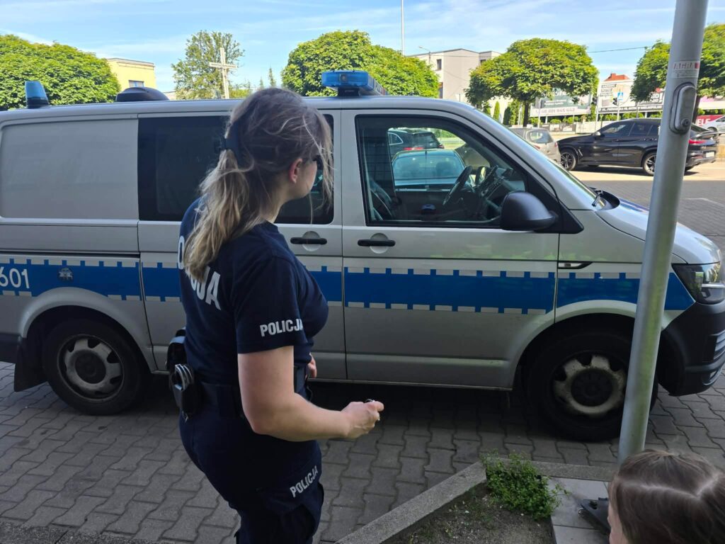 Pani policjantka w granatowym mundurze stoi na tle srebrnego wozu policyjnego.