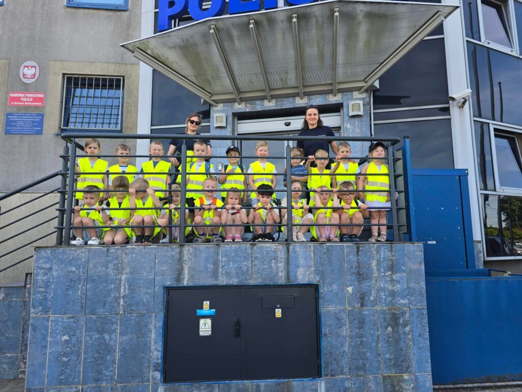 Duża grupa dzieci razem z dwójką policjantek pozują do wspólnego zdjęcia na tle Komendy Policji.