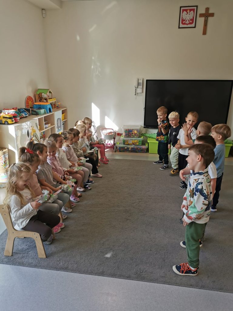 Dziewczynki siedzą na krzesłach ustawionych w rzędzie, chłopcy stoją naprzeciw