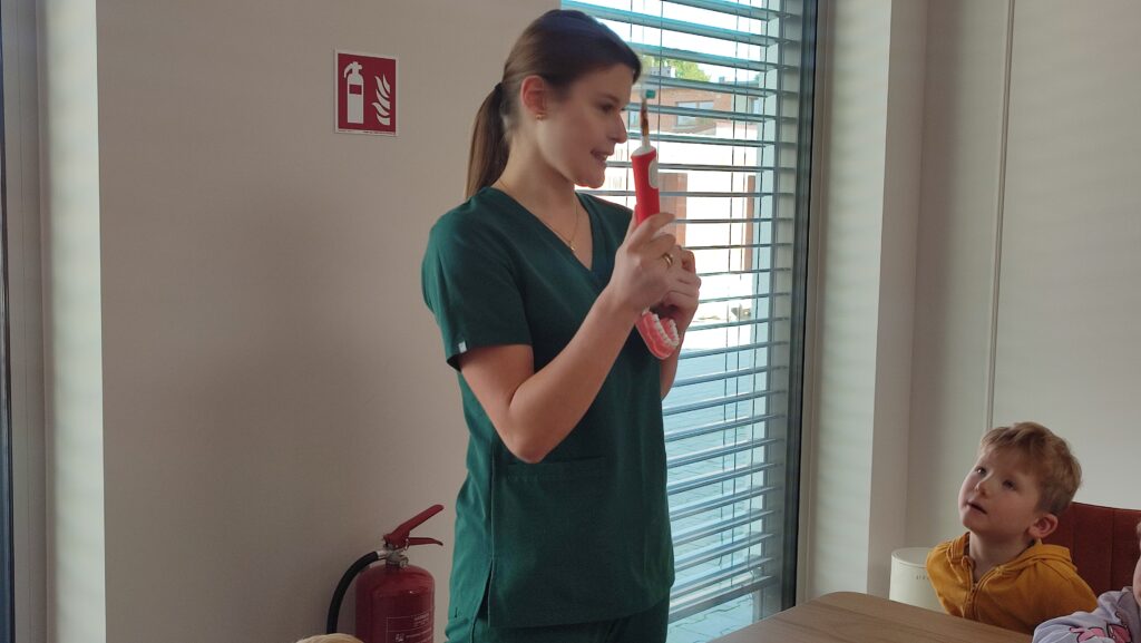 Pani dentystka w zielonym stroju pokazuje dzieciom szczotkę, pastę i jak należy szczotkować zęby.