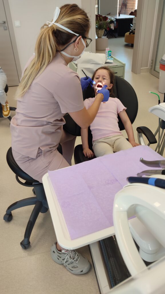 Dziewczynka siedzi na fotelu dentystycznym z otwartą buzią. Pani dentystka ogląda zęby.