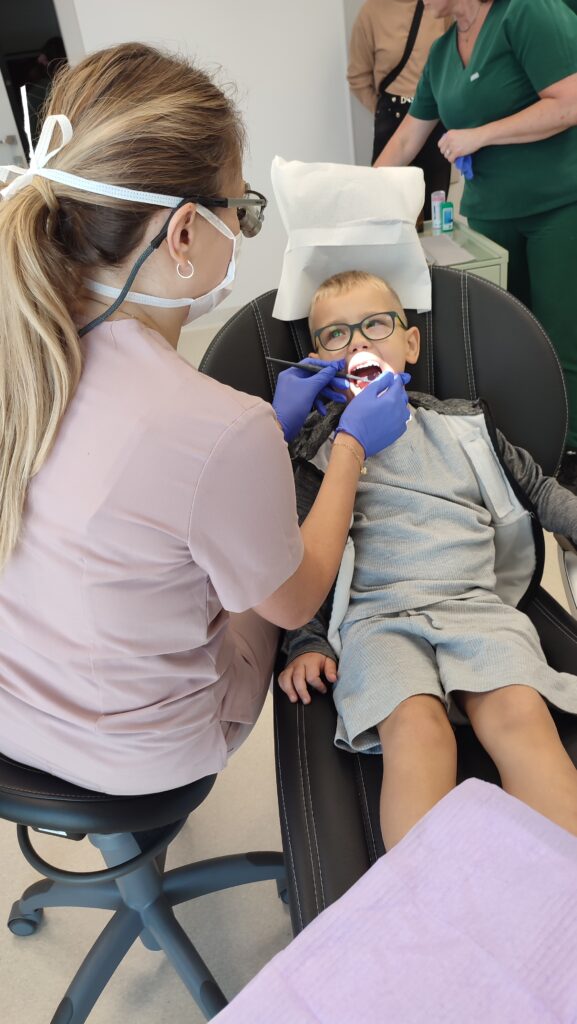 Chłopiec siedzi na fotelu dentystycznym z otwartą buzią. Pani dentystka ogląda zęby.